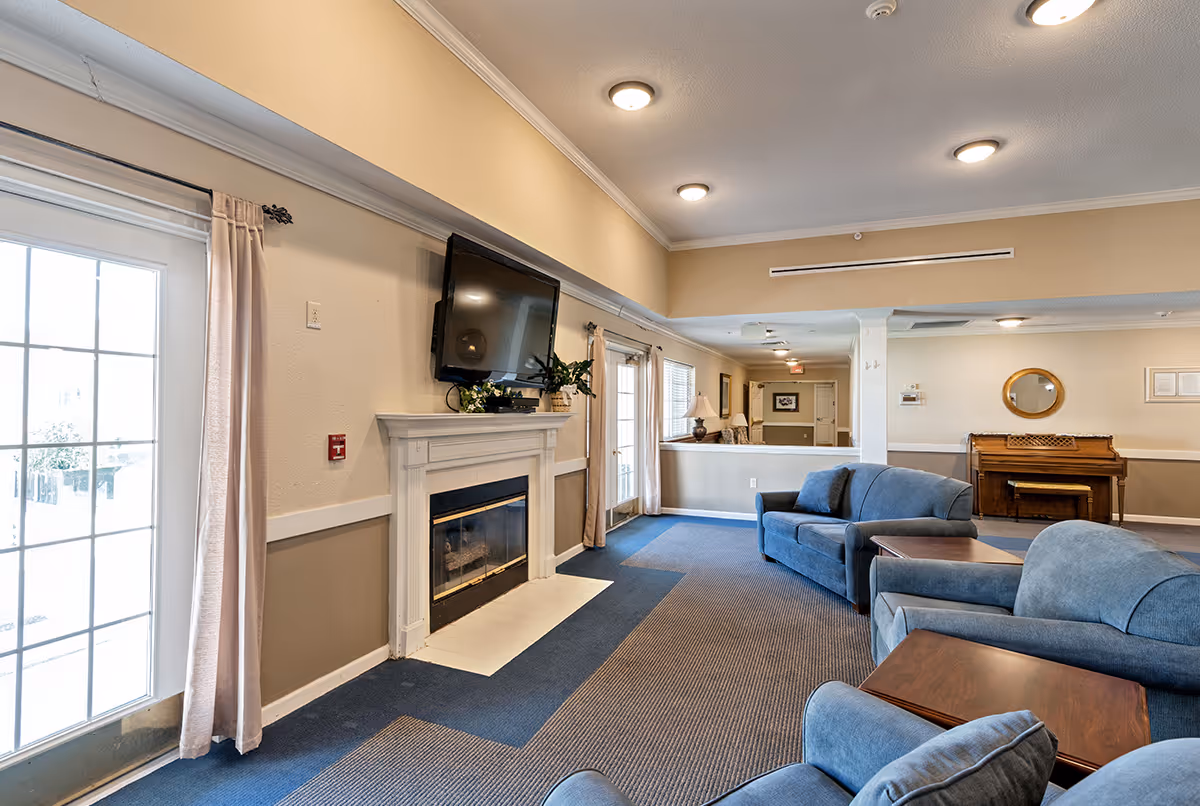 A senior living facility common area with blue upholstered sofas and armchairs arranged around wooden side tables. A white fireplace with a mounted flat-screen TV above it is on the left wall. Large windows and glass doors with beige curtains allow natural light into the room. In the background, there is a wooden piano against the wall with a round mirror above it. The room has beige walls, recessed ceiling lights, and a blue and gray carpeted floor.