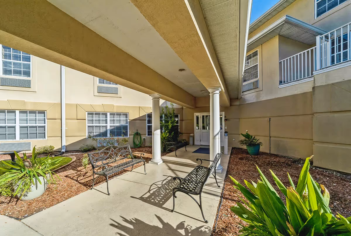 Covered exterior entrance to a senior living facility with decorative benches, columns, potted plants, and double doors.