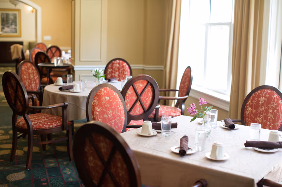 Sunlit dining room with round tables set with cups, glasses, napkins, and red upholstered chairs.