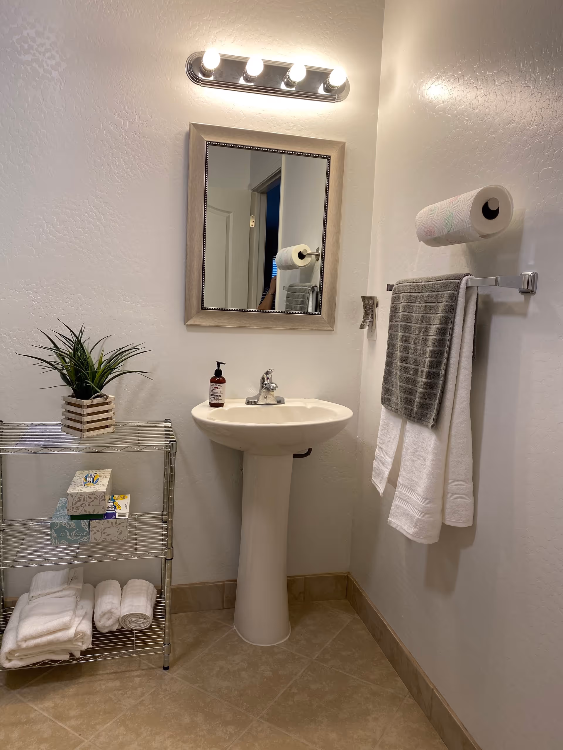 A small bathroom corner with a white pedestal sink, a rectangular mirror with a beige frame above it, and a light fixture with four bulbs mounted on the wall. To the right of the sink, a towel rack holds a gray and a white towel, with a roll of paper towels mounted above it. To the left of the sink, a metal shelving unit holds a small potted plant on the top shelf, tissue boxes on the middle shelf, and folded white towels on the bottom shelf. The floor is tiled in a beige color.