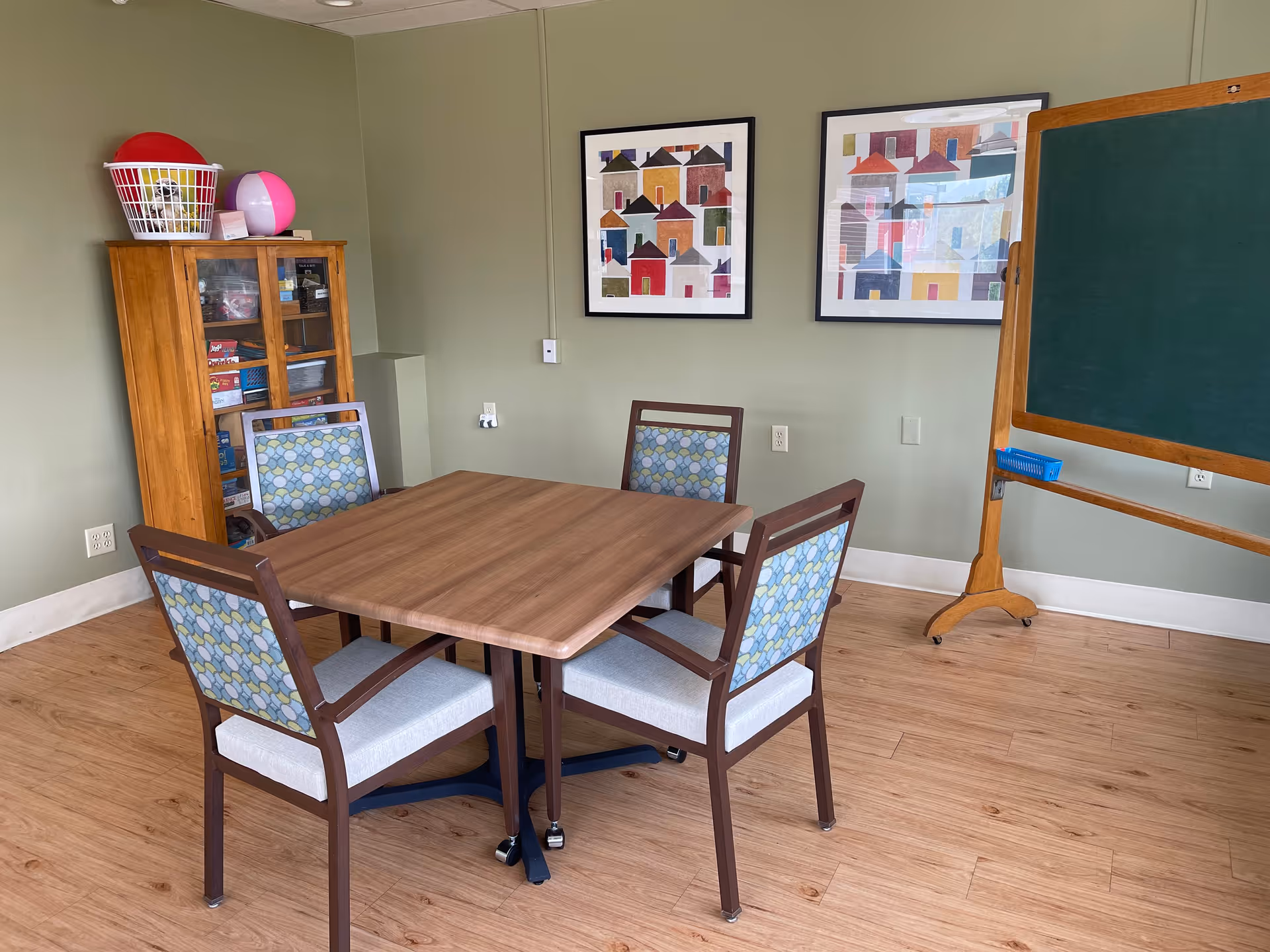 A small room with a wooden table surrounded by four chairs with patterned backs and light gray seats. On the left side, there is a wooden cabinet with glass doors containing board games and other items, with a laundry basket and a beach ball on top. Two colorful framed artworks featuring abstract house designs hang on the green wall. On the right side, there is a large green chalkboard on a wooden stand. The floor is light wood.