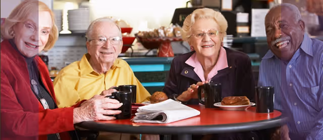 Four elderly people sitting around a red table in a dining area, smiling and holding black mugs. There are plates with pastries and a folded newspaper on the table. The background shows shelves with plates and other kitchen items.
