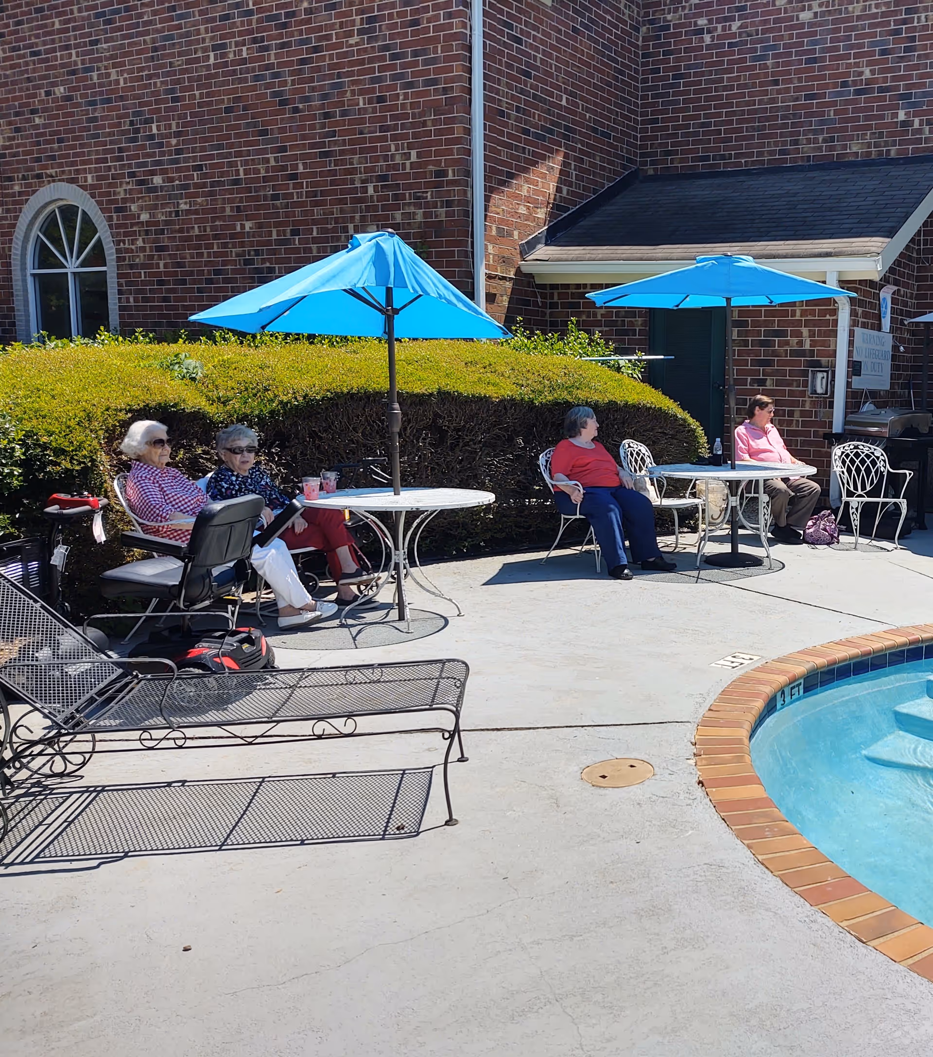 Four elderly women sitting outdoors near a swimming pool at City Center Village Senior Community. They are seated at two round tables with blue umbrellas providing shade. The background features a brick building and trimmed green bushes.