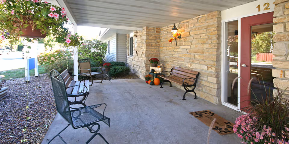 Covered outdoor patio area with metal and wooden benches, chairs, potted plants, flowers, and pumpkins near the entrance door of a building with stone walls.
