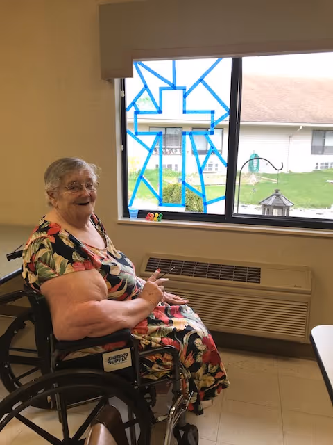 An elderly woman in a floral dress sitting in a wheelchair inside a room, smiling and pointing towards a window with a blue tape design in the shape of a cross. Outside the window, a lawn and part of a building are visible.