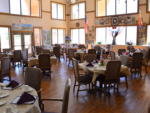 A spacious dining room with multiple round and rectangular tables covered with beige tablecloths, each set with white dishes, cups, and folded dark purple napkins. The room has large windows letting in natural light, wooden floors, and walls decorated with framed pictures and two American flags.