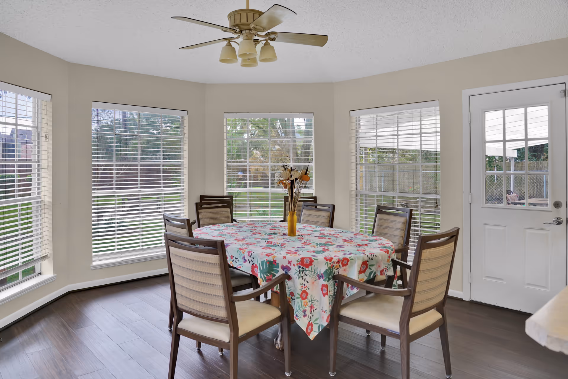 A bright dining area with a round table covered by a floral tablecloth surrounded by six wooden chairs with cushioned seats. The room has large windows with white blinds letting in natural light, a ceiling fan with lights, and a white door leading outside. Outside, a green lawn and trees are visible.