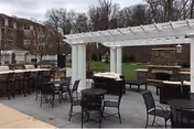 Outdoor patio with tables and chairs under a white pergola, bar seating, and an outdoor fireplace with a brick building and lawn in the background.