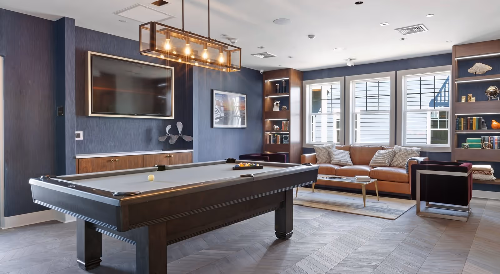 A modern living room with a pool table in the foreground, a large flat-screen TV mounted on a dark blue wall, a brown leather sofa with patterned pillows, two armchairs, a coffee table, and built-in shelves with books and decorative items. Large windows allow natural light to fill the room.