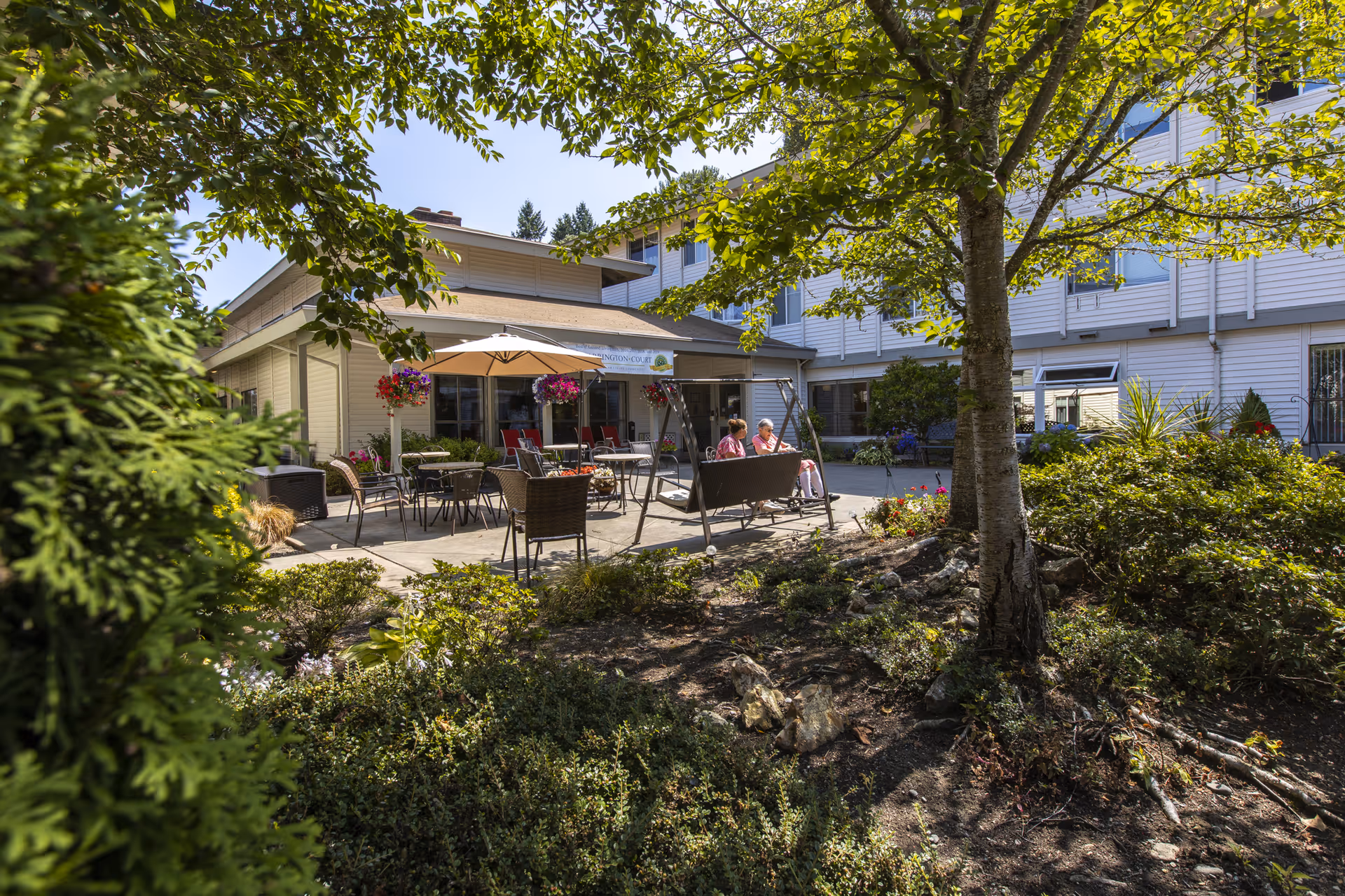 Outdoor patio area at a senior living facility with two elderly women sitting on a swing under a tree. The patio has tables, chairs, and an umbrella surrounded by greenery and flowers. The building is visible in the background under a clear blue sky.