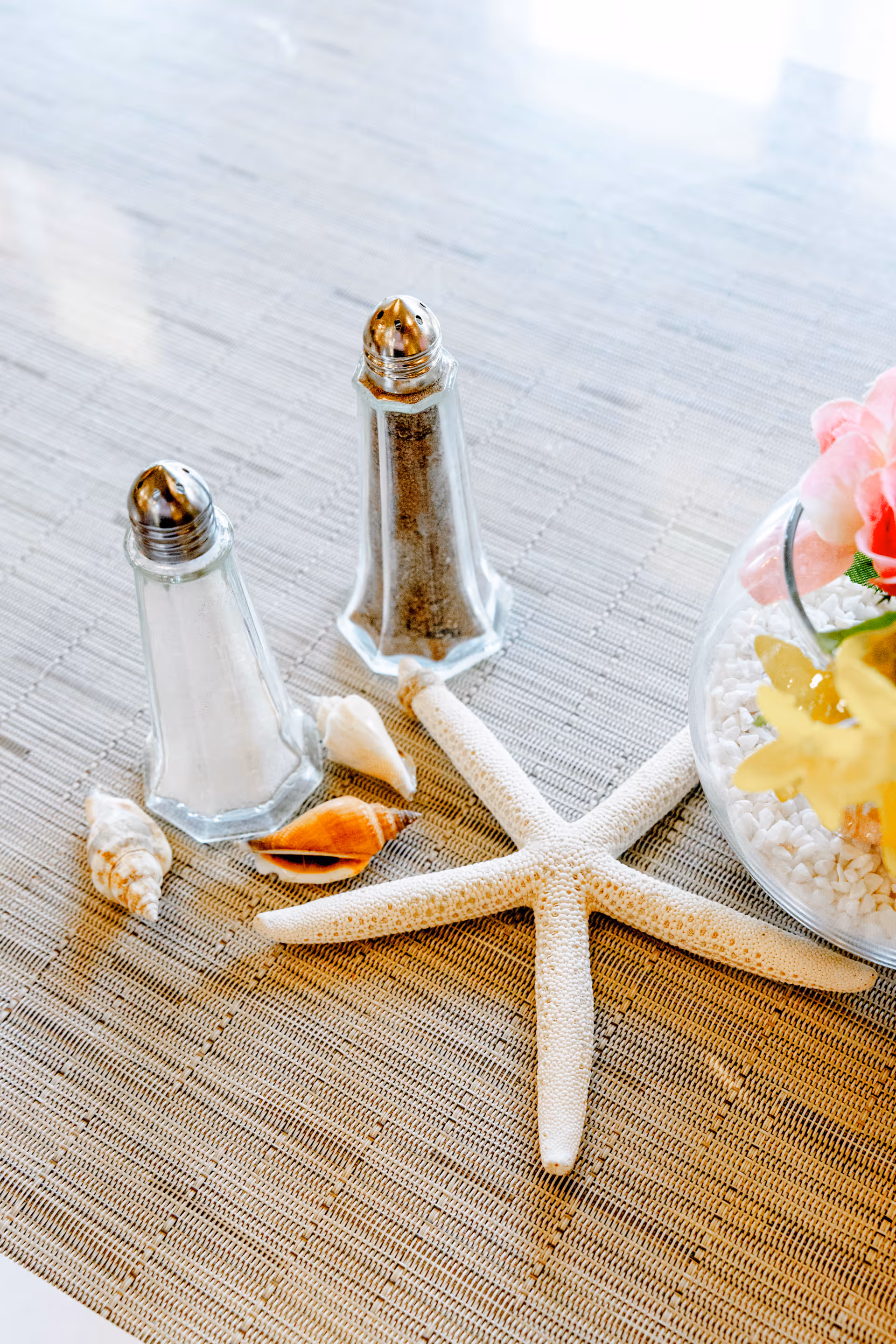 A close-up of a table setting featuring a salt shaker, a pepper shaker, a starfish, several seashells, and a glass bowl with white pebbles and colorful flowers on a woven placemat.