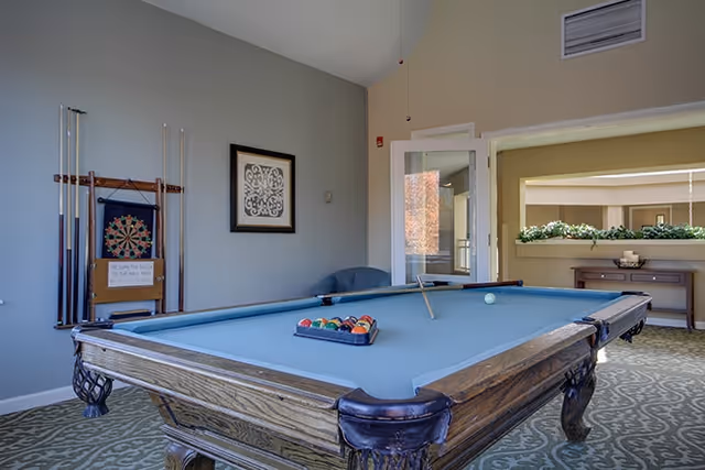 Interior room with a pool table in the center, a dartboard mounted on the wall with pool cues beside it, a framed decorative artwork on the wall, a blue chair in the corner, and a console table with greenery and candles in the background.