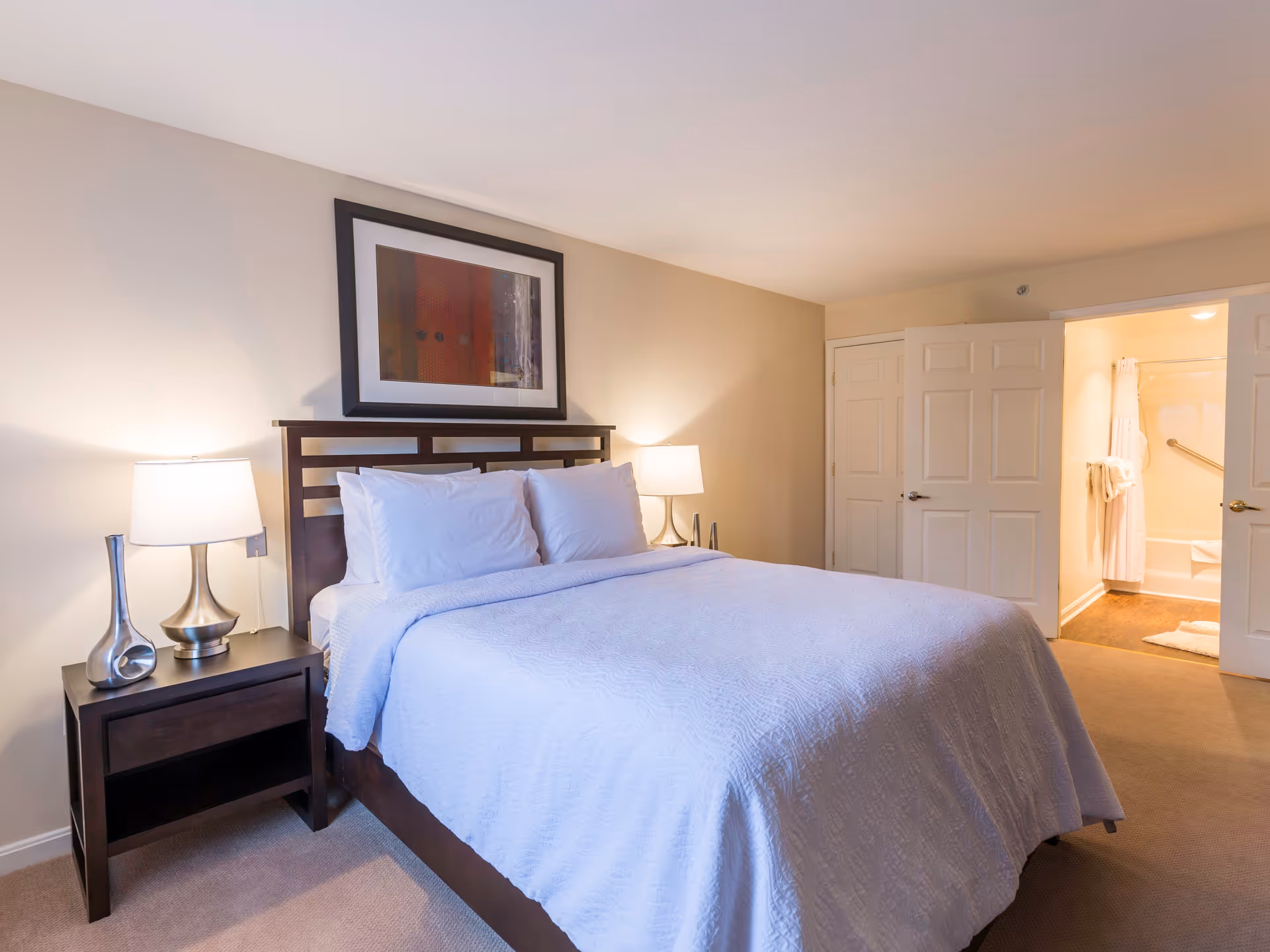 A neatly made bed with white bedding and pillows in a bedroom. There are two bedside tables with modern lamps on each side of the bed. A framed abstract artwork hangs above the headboard. In the background, an open door reveals a bathroom with a shower curtain, towel rack, and grab bar.