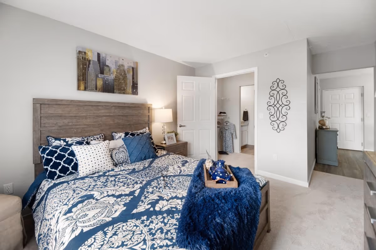 A bedroom with a wooden headboard bed dressed in blue and white patterned bedding and multiple pillows. A wooden nightstand with a lamp and picture frame is beside the bed. The room has light gray walls and carpeted floor. An open door reveals a walk-in closet and a glimpse of a bathroom. A decorative metal wall art hangs on the wall near the door, and a blue throw blanket with a wooden tray holding blue ceramic items is on the bed.
