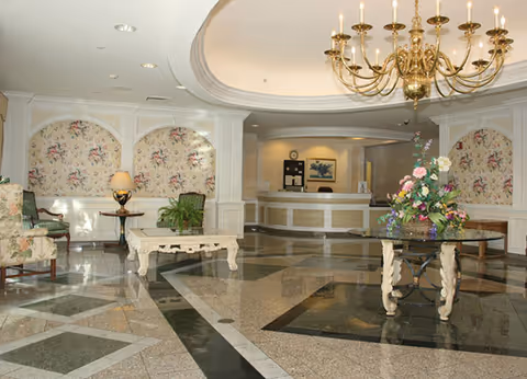 A spacious senior living facility lobby with polished marble floors featuring geometric patterns. There is a large chandelier hanging from the ceiling, floral arrangements on a round table in the center, and seating areas with floral upholstered chairs and a small table with a lamp. The reception desk is visible in the background with a clock and framed artwork on the wall.