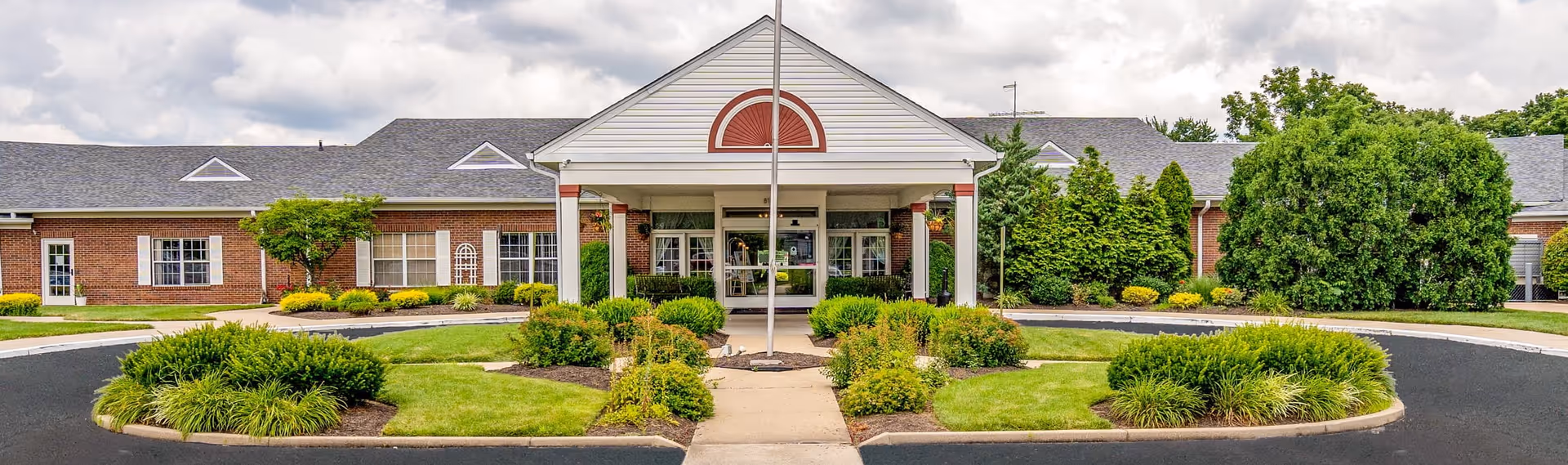 Front exterior view of The Anderson Nursing and Rehabilitation Center, featuring a single-story brick building with a covered entrance, surrounded by well-maintained landscaping and greenery under a partly cloudy sky.