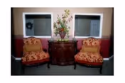 Two floral upholstered armchairs flank a wooden console with a vase of flowers beneath two windows.