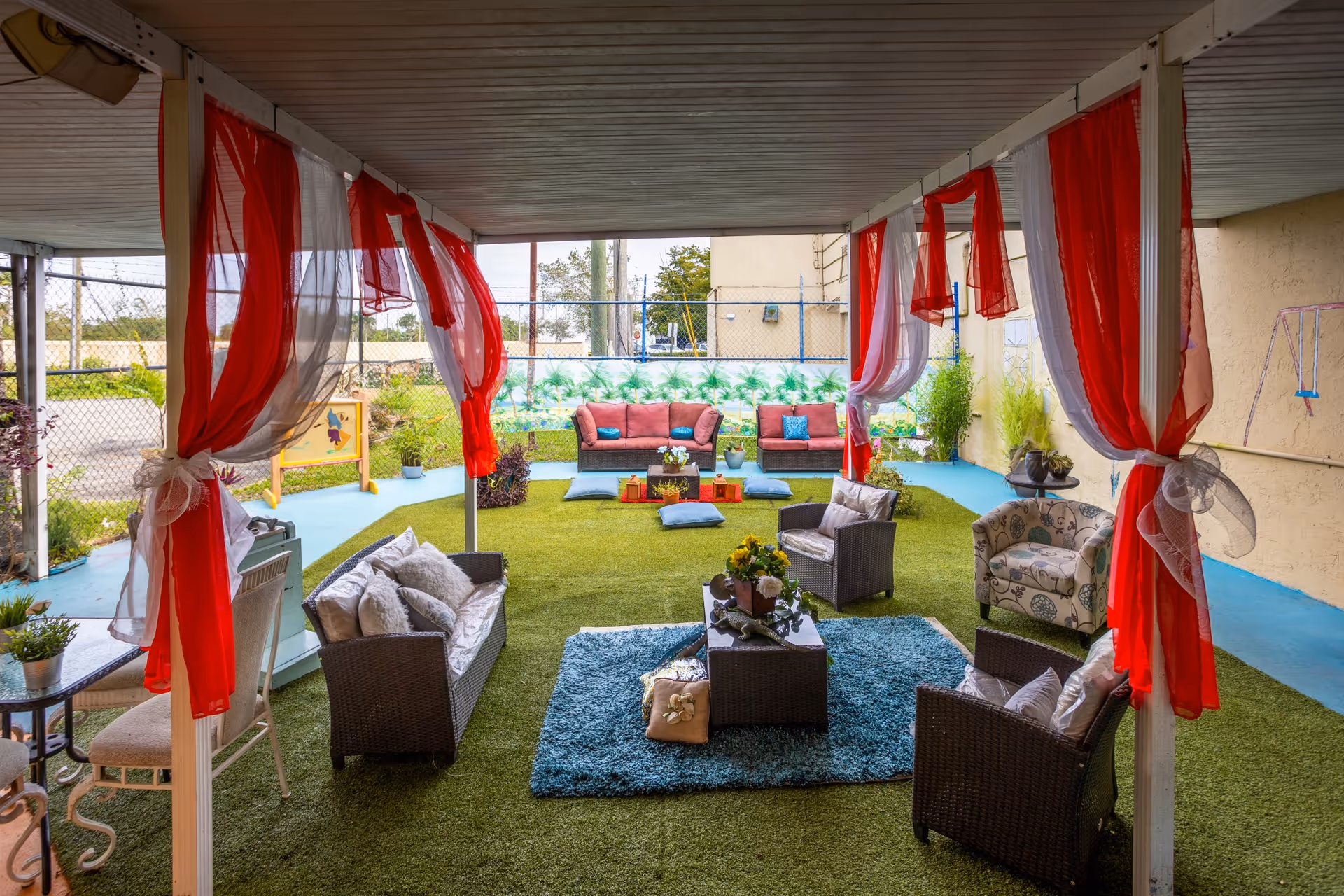 Covered outdoor seating area with artificial grass flooring, featuring multiple cushioned chairs and sofas arranged around small tables. Red and white sheer curtains are tied to the white pillars supporting the roof. In the background, there is a fenced area with a mural of palm trees and some plants, along with a swing set on the right side.