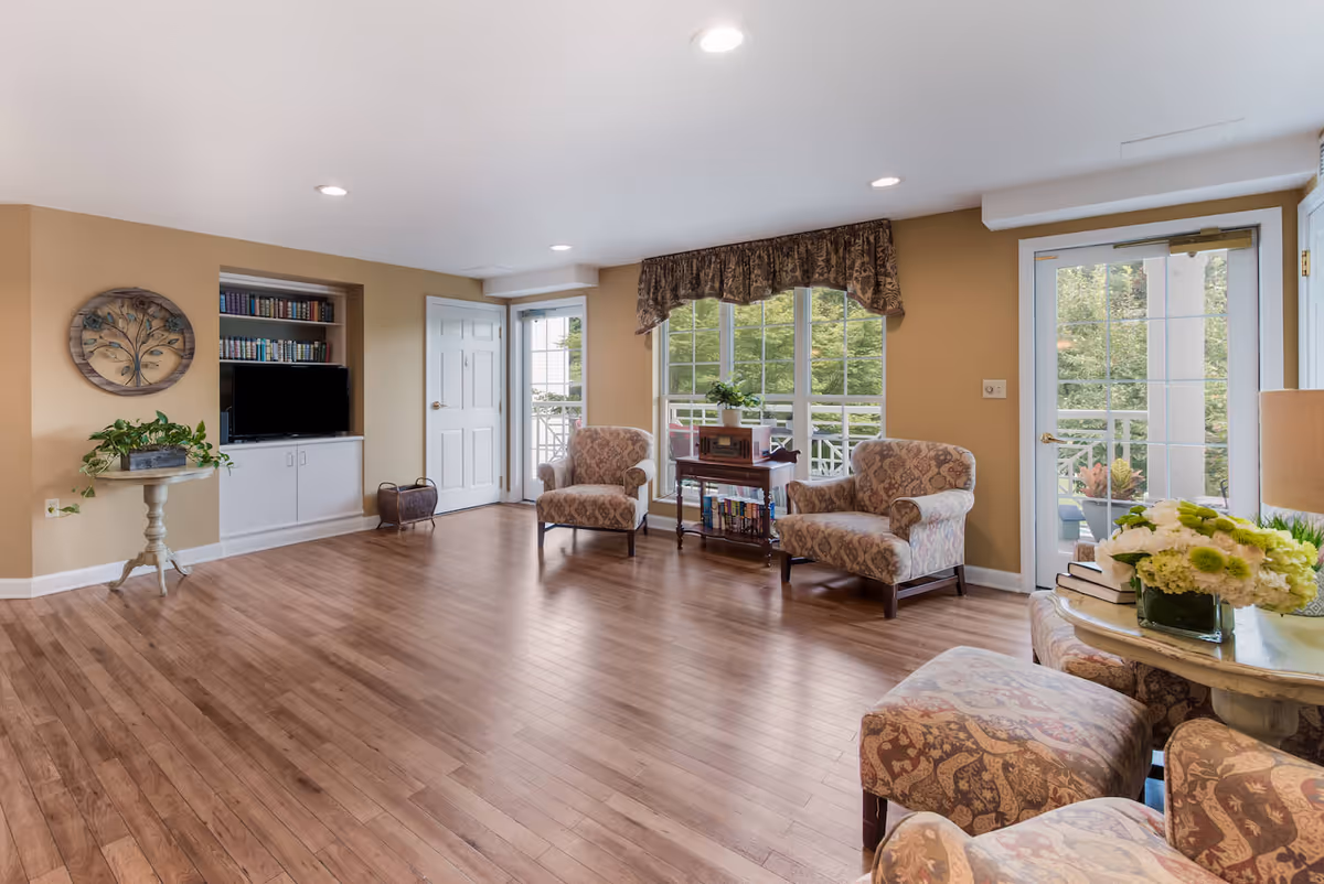 A bright and spacious living room with wooden flooring, beige walls, and recessed ceiling lights. The room features two patterned armchairs near large windows with a floral valance, a small table with books and a plant, a built-in bookshelf with a TV, and a round table with a flower arrangement and books. There are glass doors leading outside, allowing natural light to fill the space.