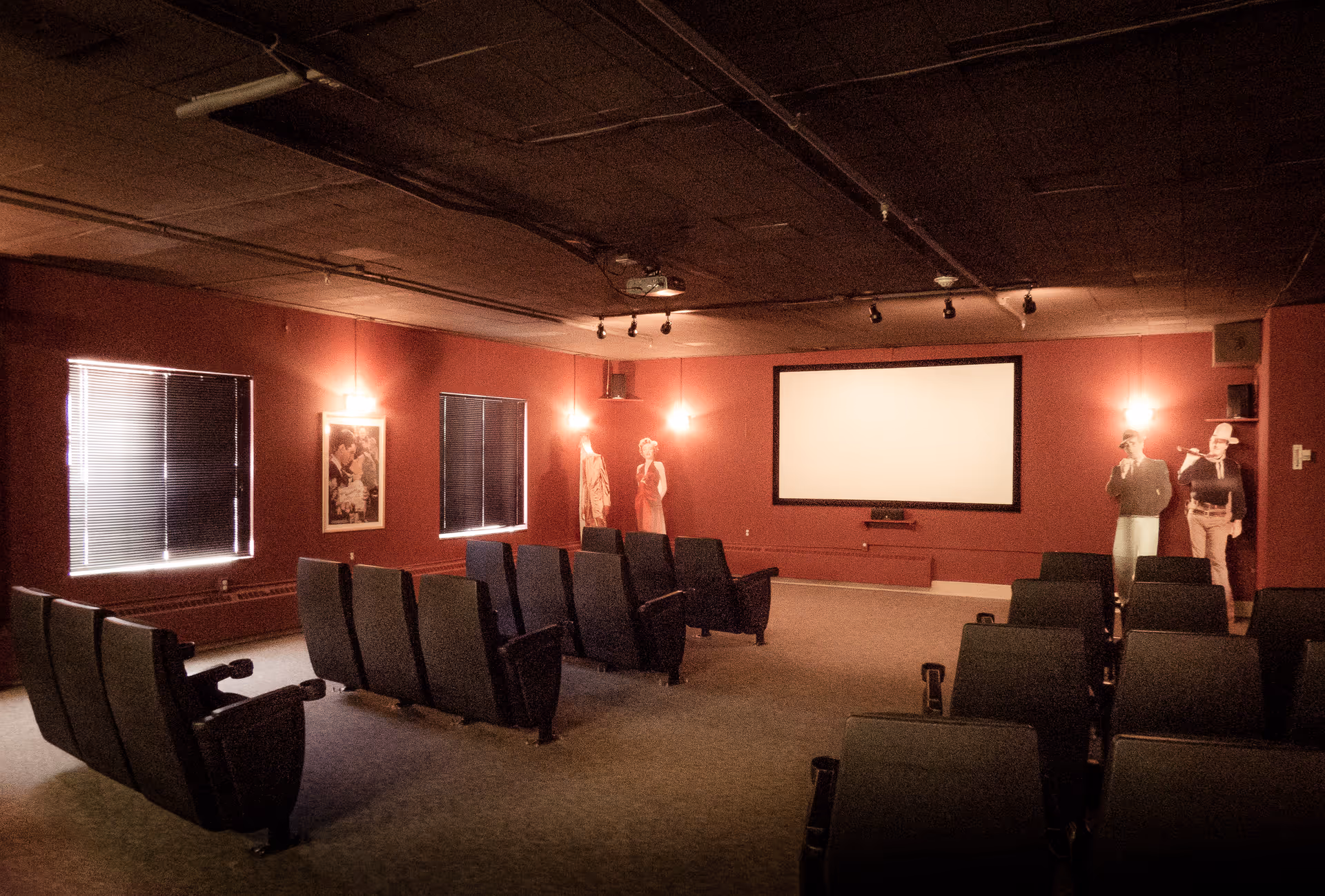 A small theater room with rows of black cushioned seats facing a large blank projection screen on a red wall. The room has dim lighting with wall sconces and two windows with closed blinds. There are life-sized cutouts of people near the screen and a framed picture on the wall.