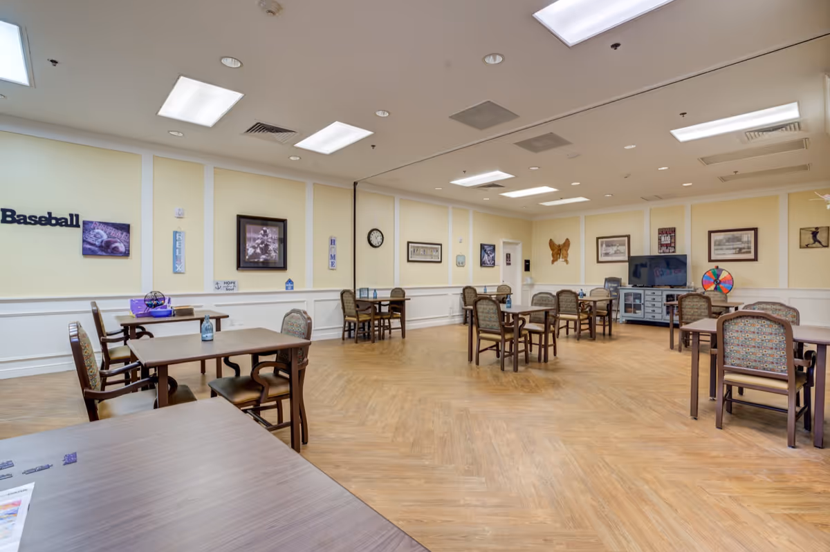 A spacious activity or common room with several tables and chairs arranged around the room. The walls are decorated with framed pictures, signs, and a large butterfly decoration. There is a television on a cabinet in the back corner and a colorful spinning wheel next to it. The room has a light wood floor and bright overhead lighting.