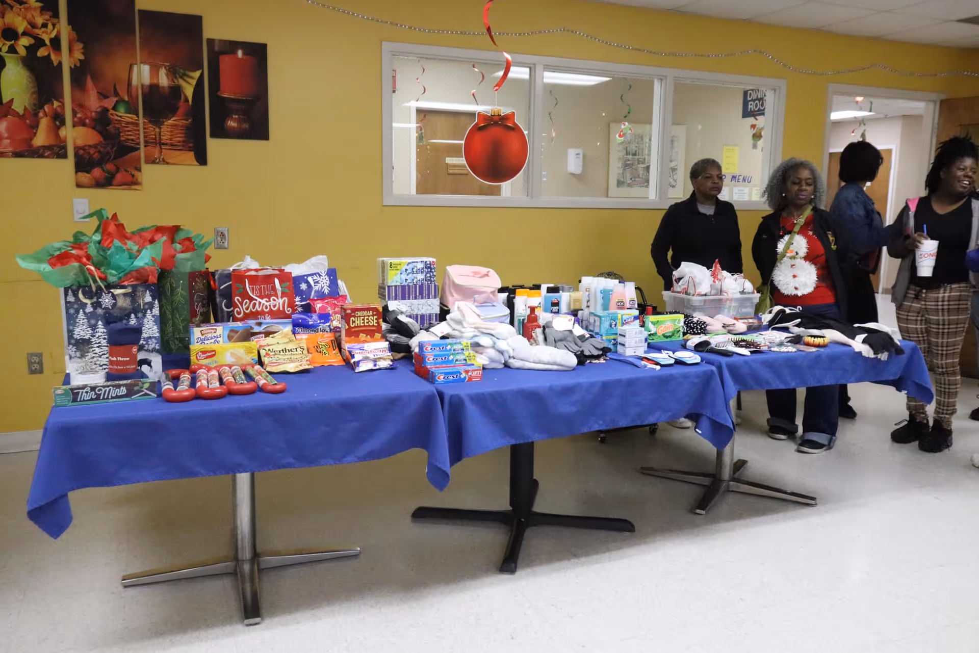 A table covered with a blue tablecloth displaying various items including snacks, toiletries, gloves, and other personal care products. Four women stand behind the table in a room with yellow walls decorated with holiday-themed pictures and hanging ornaments.