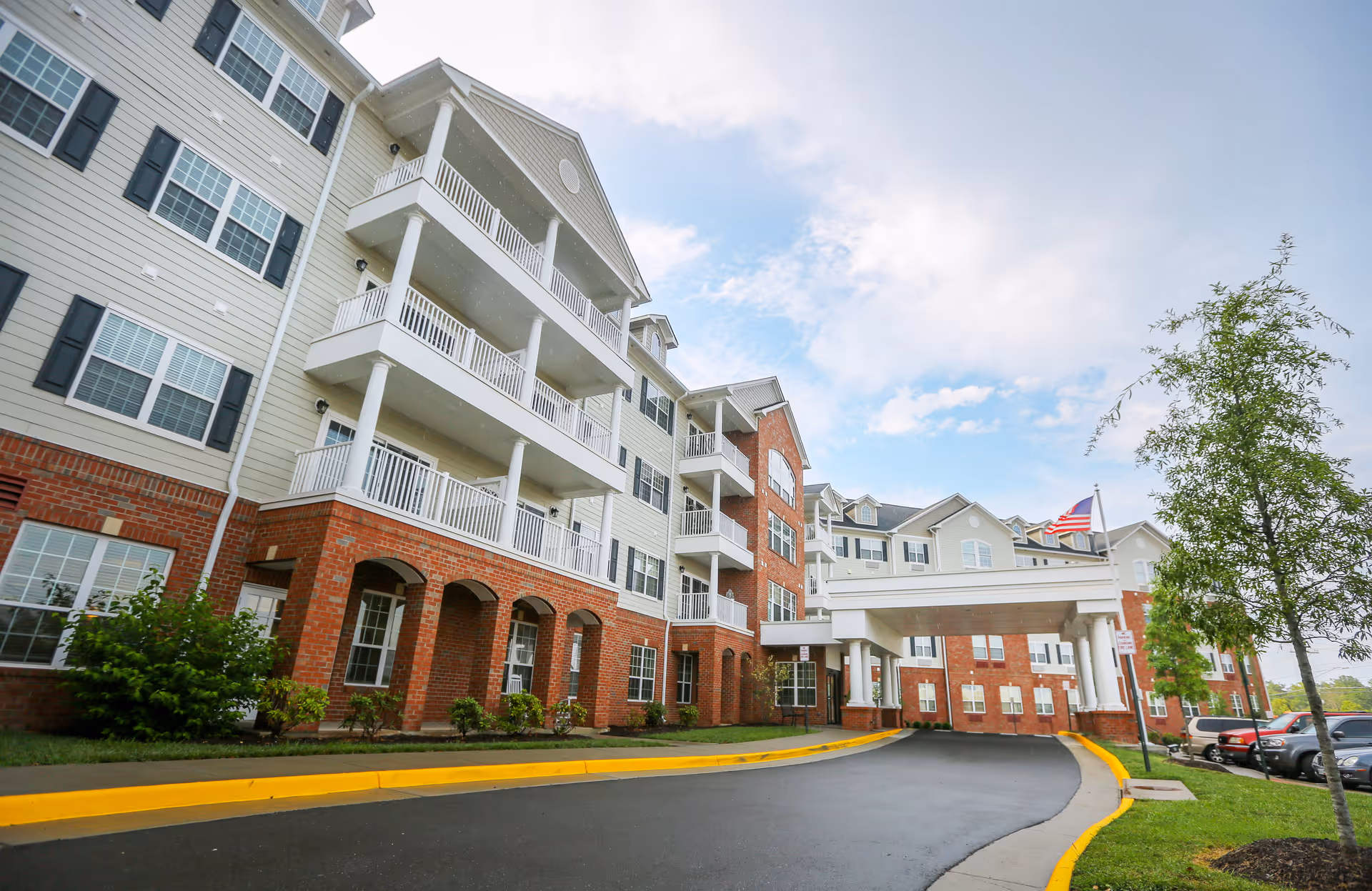 Front entrance of a multi-story senior living building with balconies, brick lower facade, a covered porte-cochere, and an American flag.