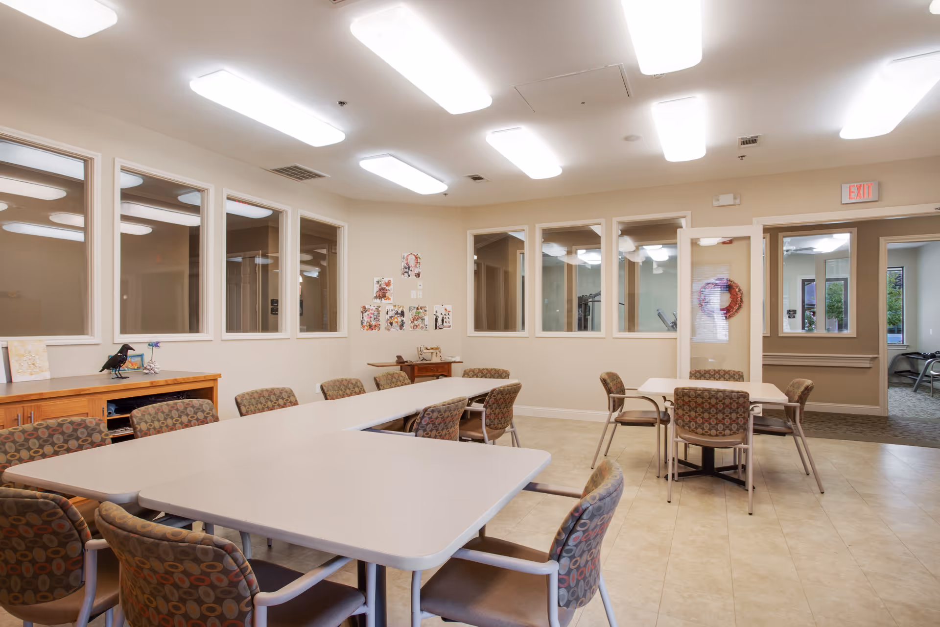 A well-lit room with multiple tables and cushioned chairs arranged for group seating. The walls have several windows looking into adjacent rooms, and there are some decorations and artwork on the walls. The floor is tiled, and there is a wooden cabinet against one wall with a decorative bird and other small items on top.