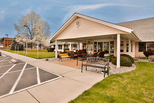 Covered front entrance of a senior living facility with benches and chairs on a paved patio, adjacent sidewalk and parking spaces.