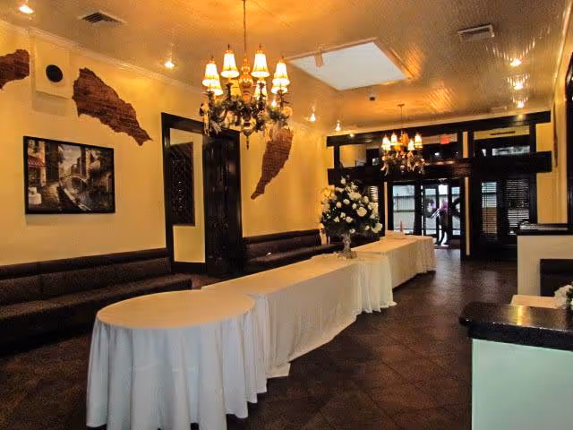 Interior view of a reception or lobby area with long tables covered in white tablecloths, floral arrangements, chandeliers hanging from the ceiling, dark cushioned benches along the walls, and a person visible near the glass entrance doors at the far end.