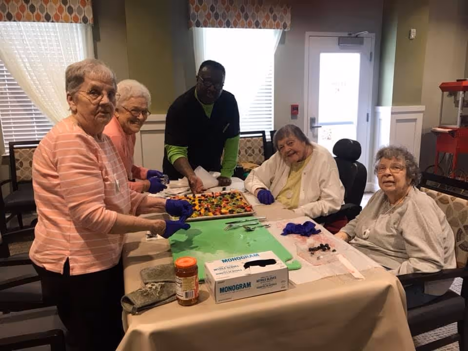 Four elderly women and one caregiver gathered around a table in a well-lit room, preparing or enjoying a colorful fruit tray. The women are seated and standing, some wearing gloves, with a box of disposable gloves and a jar on the table. The room has windows with patterned valances and a door in the background.