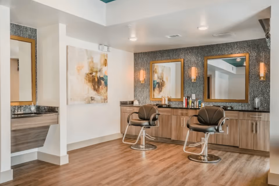 Interior view of a salon area with two black salon chairs in front of a wooden cabinet with a black countertop. Two large mirrors with wooden frames are mounted on a textured wall above the cabinet, flanked by wall-mounted light fixtures. A piece of abstract art hangs on a white wall to the left, and a small sink with a mirror is visible on the far left side. The floor is wood, and the ceiling has recessed lighting.