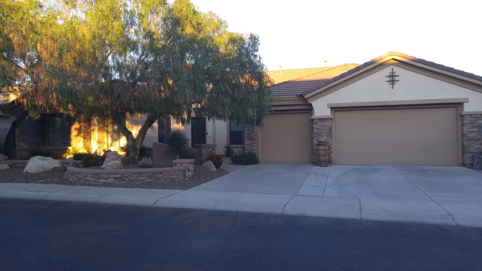 Exterior view of a single-story assisted living care home with a two-car garage, stone accents on the walls, a tree and landscaped area with rocks and plants in front, and a paved driveway and street.