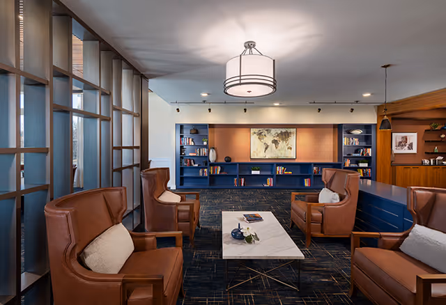 A cozy senior living common area with four brown leather armchairs arranged around a rectangular white marble coffee table. The room features a dark carpet with a subtle pattern, blue built-in bookshelves filled with books and decorative items, and a world map artwork on the back wall. A modern ceiling light fixture illuminates the space, and there is a wooden partition with shelves on the right side.