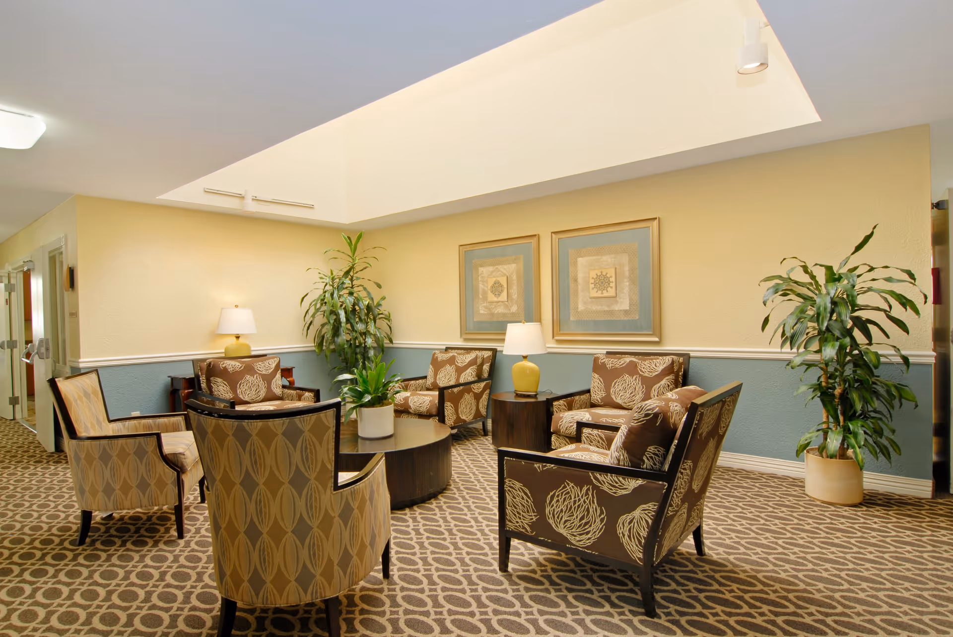 A cozy seating area in a senior living facility with six patterned armchairs arranged around a round wooden coffee table. The room has a beige and blue color scheme with two framed artworks on the wall, two table lamps with yellow bases, and two large potted plants. The ceiling features a skylight allowing natural light to brighten the space.