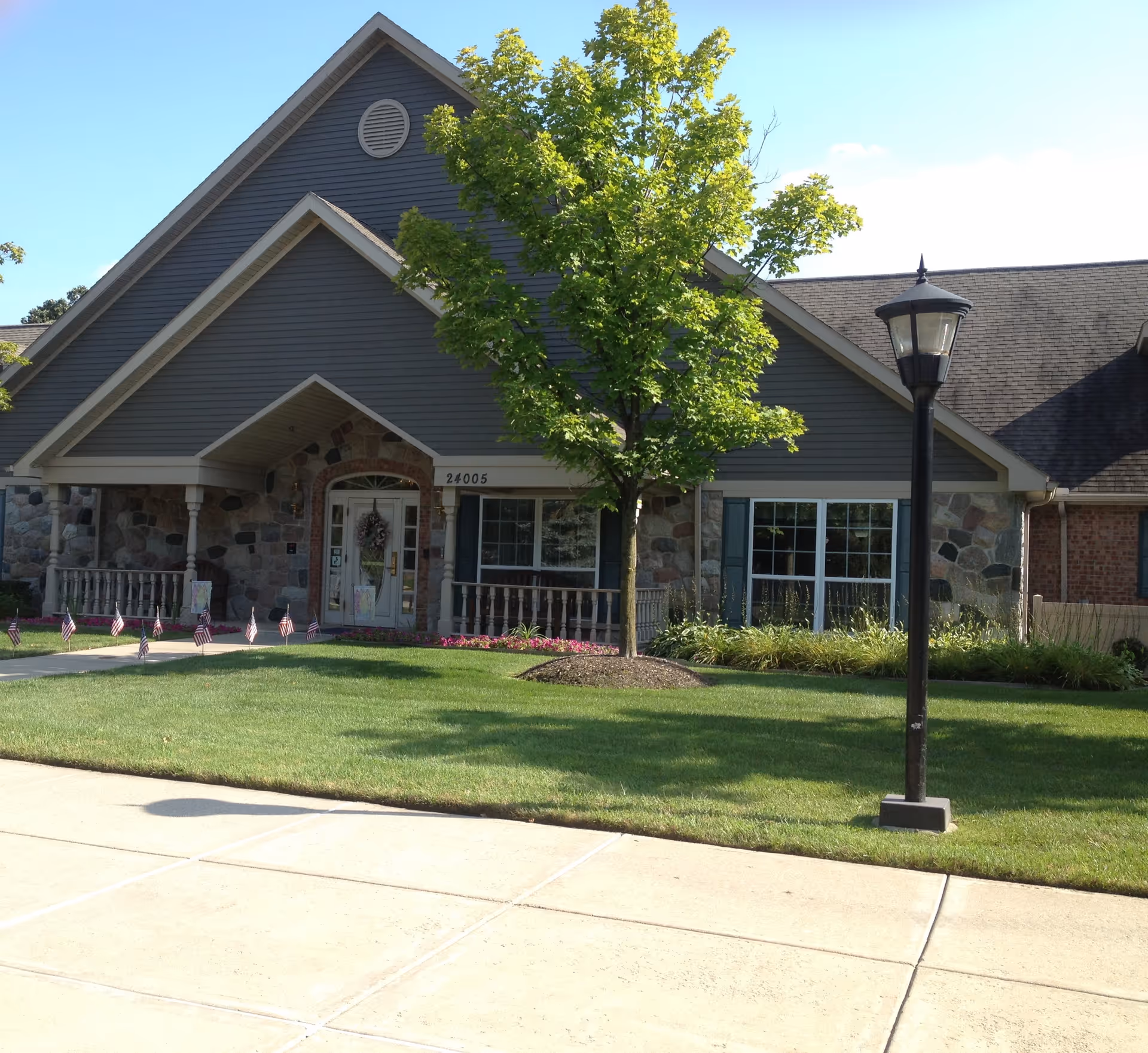 Exterior view of a single-story building with a peaked roof, stone and siding facade, a front porch with columns, a tree in the lawn, and a black street lamp near the sidewalk.