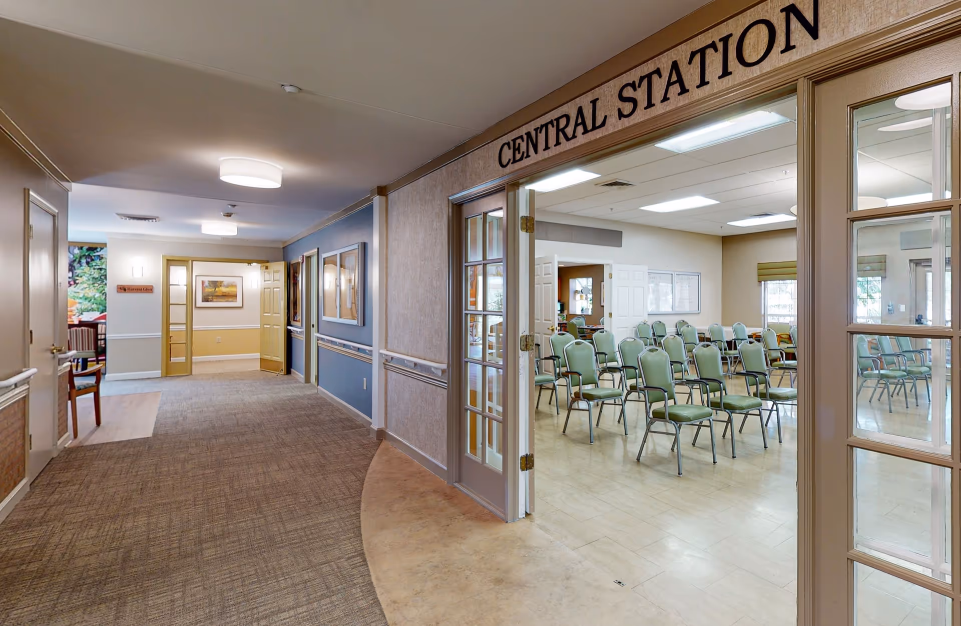Interior view of a senior living facility hallway leading to a room labeled 'Central Station' with multiple green chairs arranged in rows inside. The hallway has carpeted flooring and walls decorated with framed pictures and a sign reading 'Harvest Glen'.