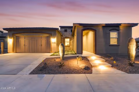 Exterior view of a modern single-story house at dusk with warm lighting illuminating the entrance and garage. The front yard features desert landscaping with cacti and rocks, and a concrete driveway and walkway leading to the front door.