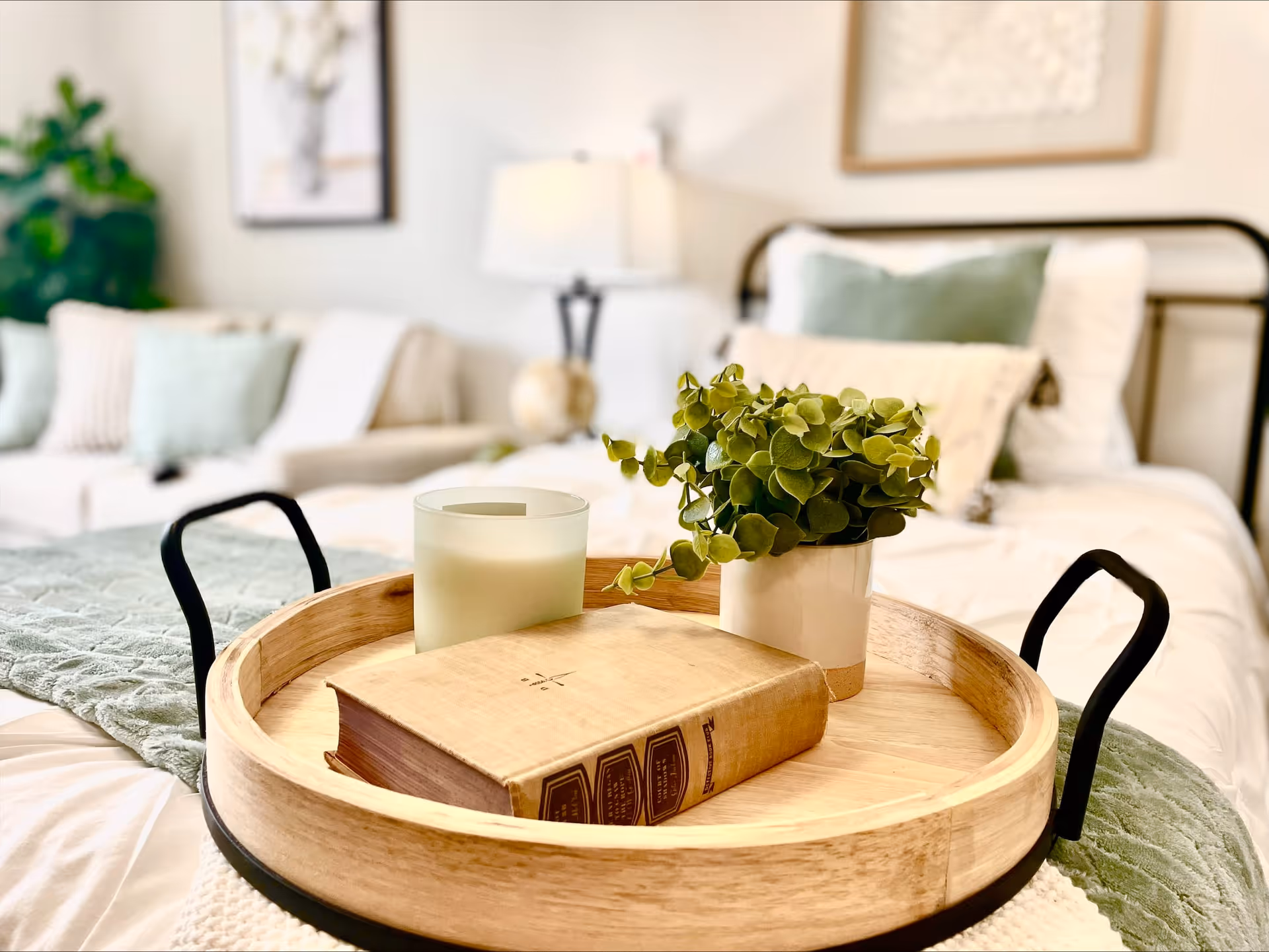 A close-up of a wooden tray on a bed, holding a green potted plant, a closed book, and a candle. In the background, a bed with white and green pillows and a soft blanket is visible, along with a lamp and framed artwork on the wall.