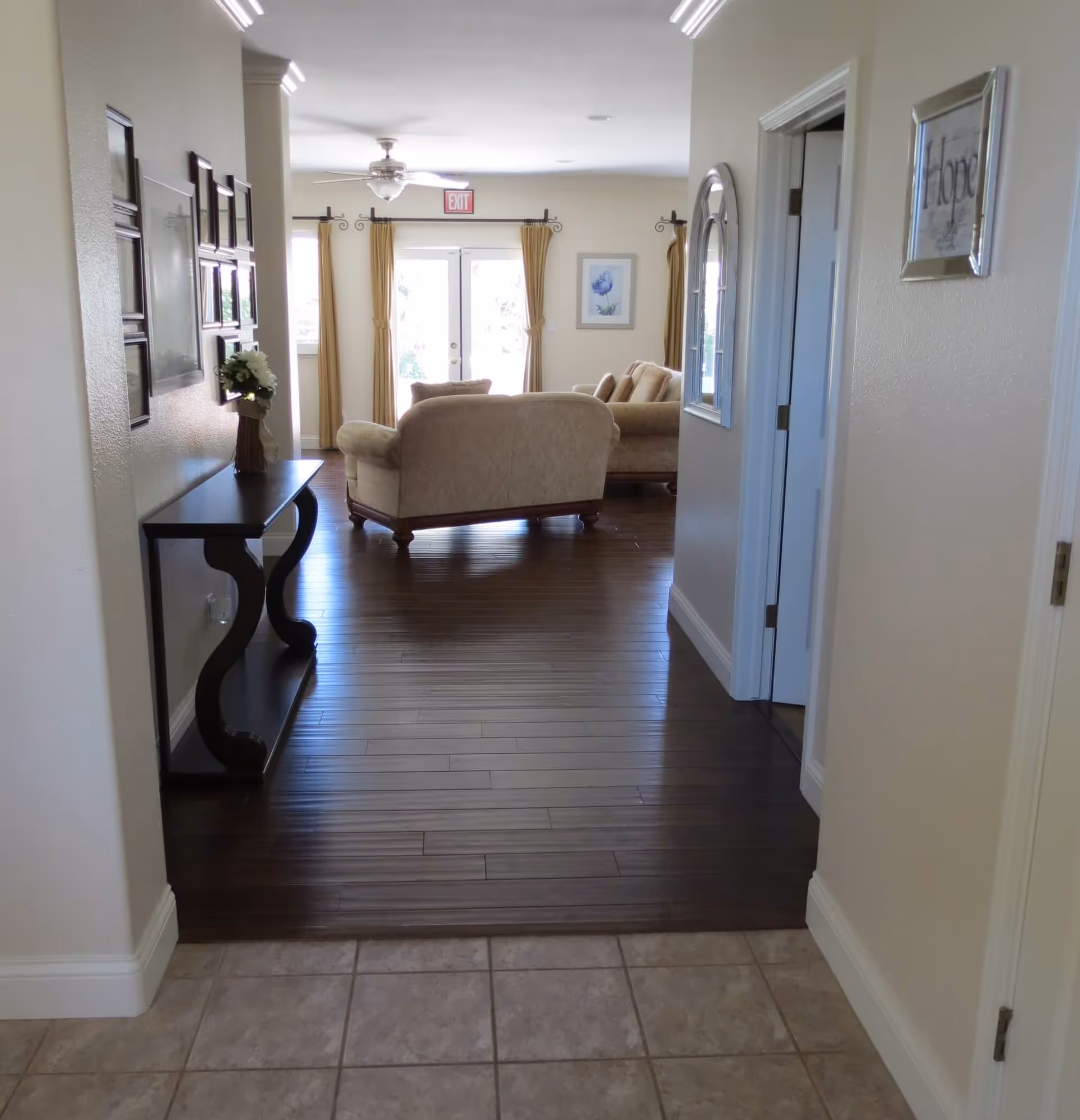 View down a hallway with tiled floor leading to a living room area with wooden flooring, beige sofas, a ceiling fan, and large windows with curtains. A black console table with a flower vase is on the left side of the hallway, and framed pictures and a mirror are on the walls.