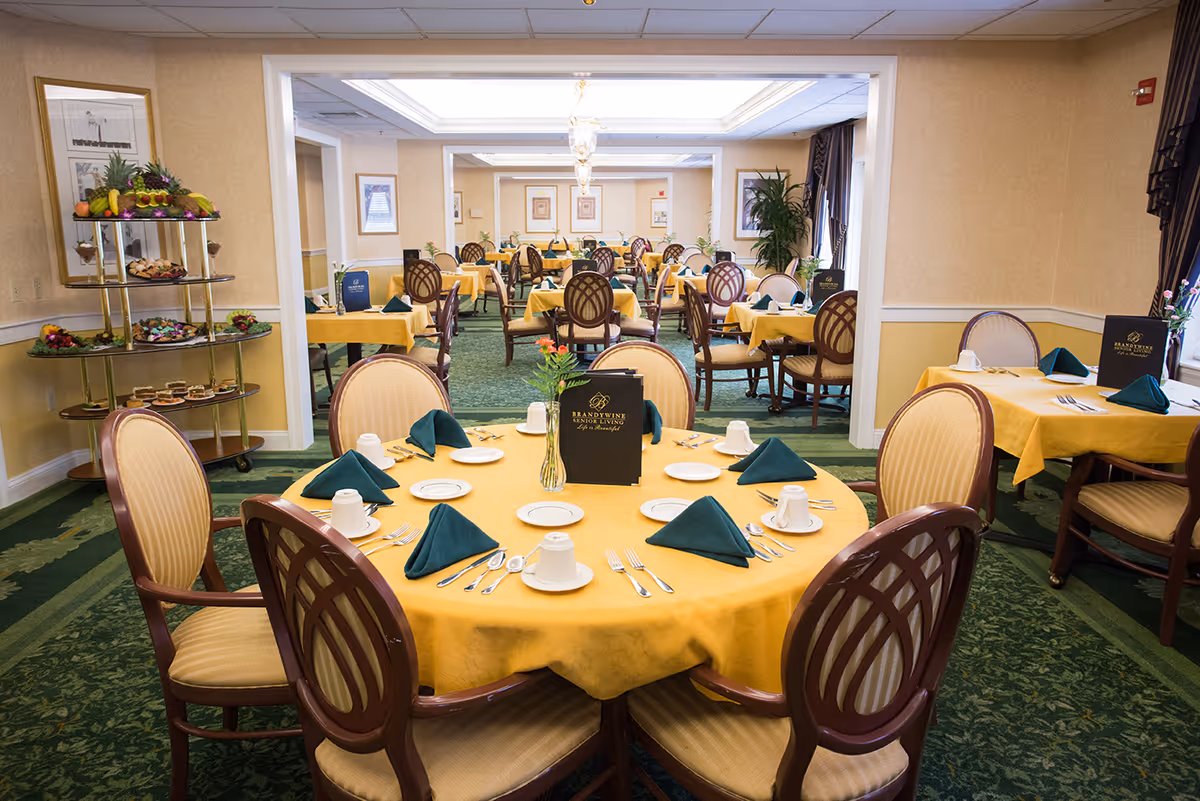 A spacious dining room with multiple round and square tables covered with yellow tablecloths and set with white plates, cups, silverware, and green folded napkins. The chairs have wooden frames with beige cushioned seats and backs. A three-tiered serving cart with various fruits and desserts is visible on the left side. The room has green carpet with a floral pattern, beige walls, framed artwork, and large windows with dark curtains.