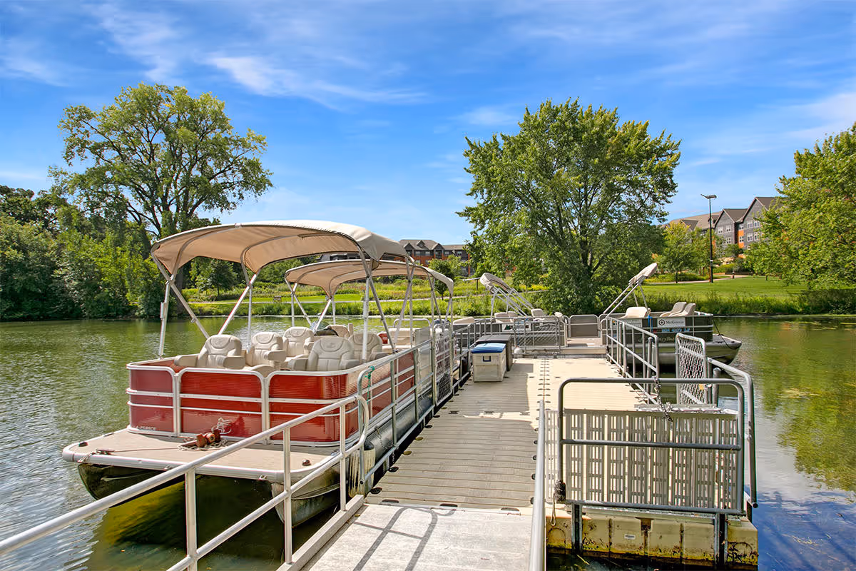A dock extending into a calm lake with two pontoon boats moored on either side. The boats have beige seats and canopy covers. Surrounding the lake are green trees and grass, with a few buildings visible in the background under a clear blue sky.