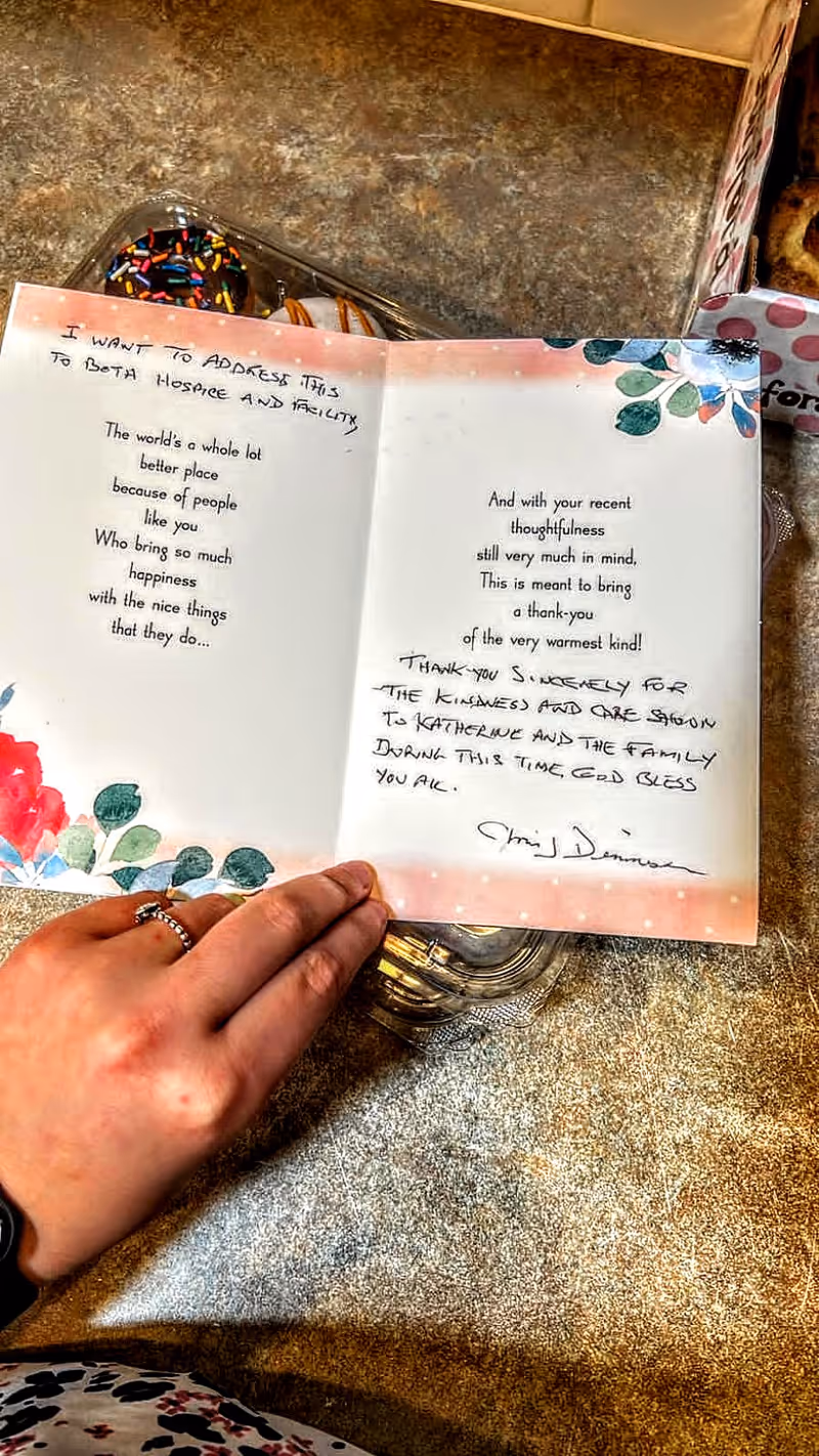 A person holding an open thank-you card with handwritten messages inside. The card has floral decorations on the corners and is placed on a countertop with a container of donuts partially visible in the background.