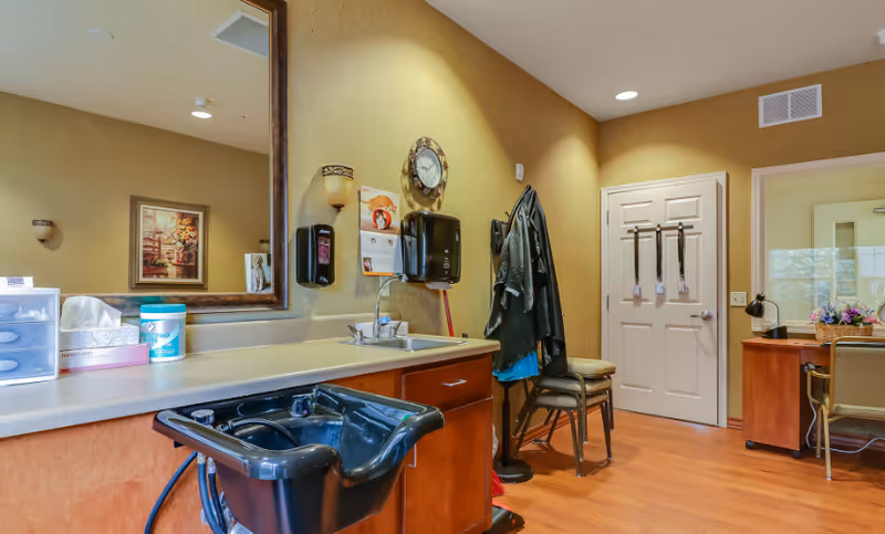 Interior view of a room in Briarwood Assisted Living & Memory Care featuring a black salon-style sink with faucet, countertop with tissue box and cleaning supplies, a large wall mirror, a clock, wall-mounted soap and paper towel dispensers, a coat rack with hanging garments, chairs, and a desk with a lamp and flower arrangement near a window.