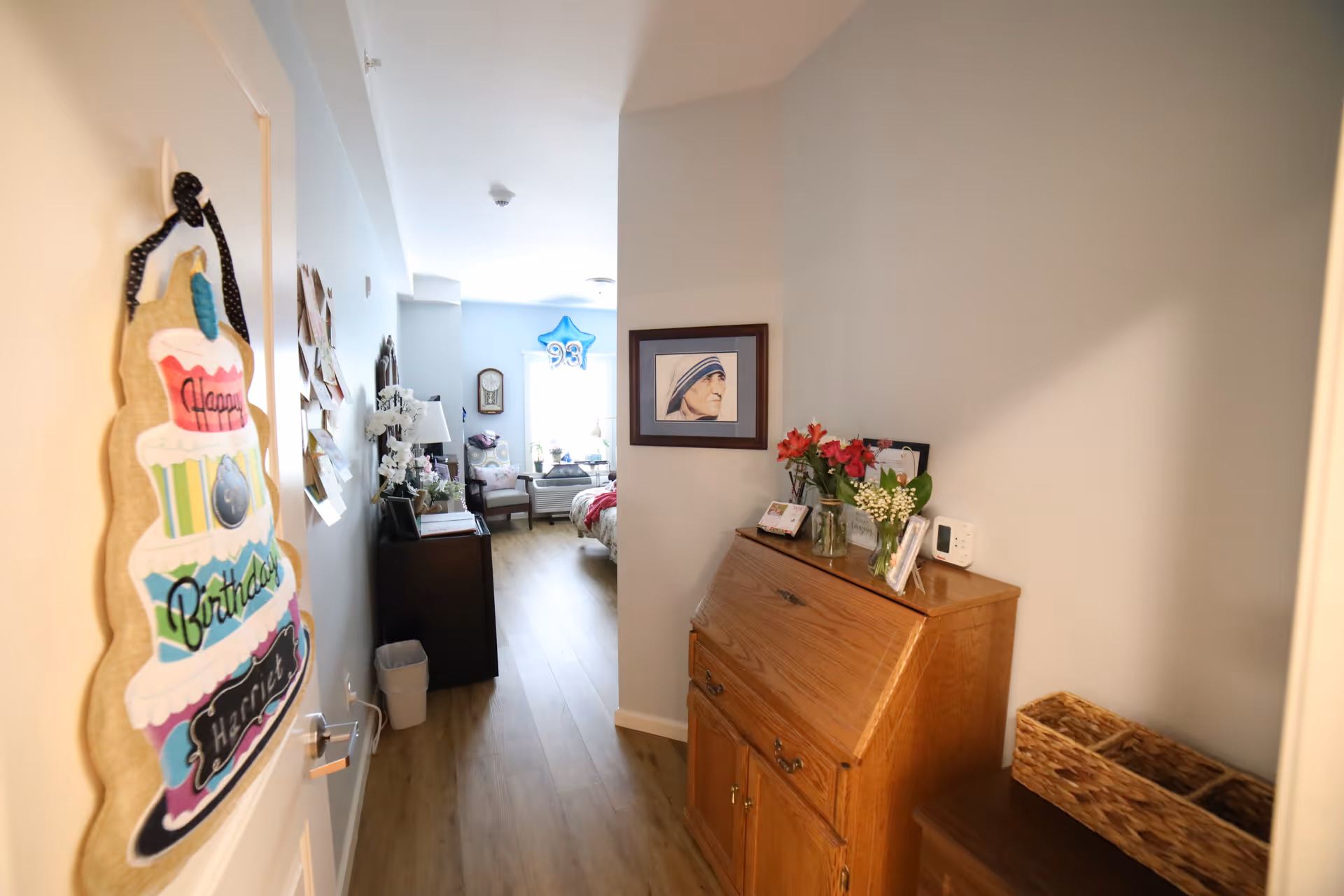 View down a hallway into a decorated bedroom with a 'Happy Birthday' sign on the door, a wooden cabinet with flowers and framed photos on the right, and a bed with a blue star-shaped balloon with the number 93 in the background.