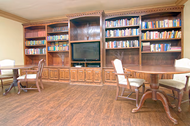 A cozy room with wooden flooring featuring a large built-in wooden bookshelf filled with books and a television in the center. There are two round wooden tables, each surrounded by cushioned chairs, positioned on either side of the room.