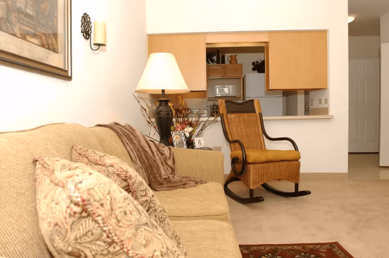 Cozy living room with a beige sofa and patterned pillows, a floor lamp, and a wooden rocking chair facing a kitchen pass-through.