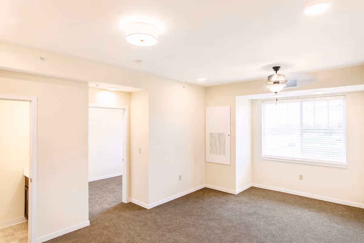 Empty room with beige walls and brown carpet, featuring a window with blinds, a ceiling fan with light, and two doorways leading to adjacent rooms.