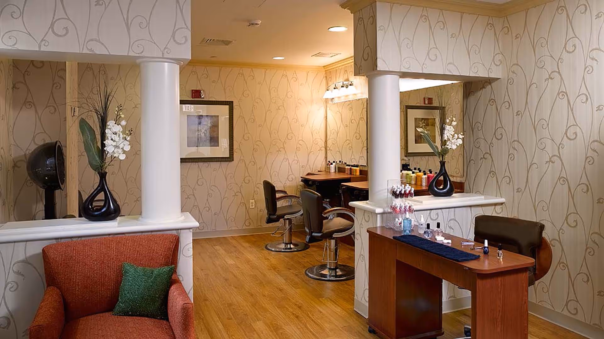 Interior salon area with styling chairs, a manicure table, decorative columns and a cushioned armchair in a senior living facility.