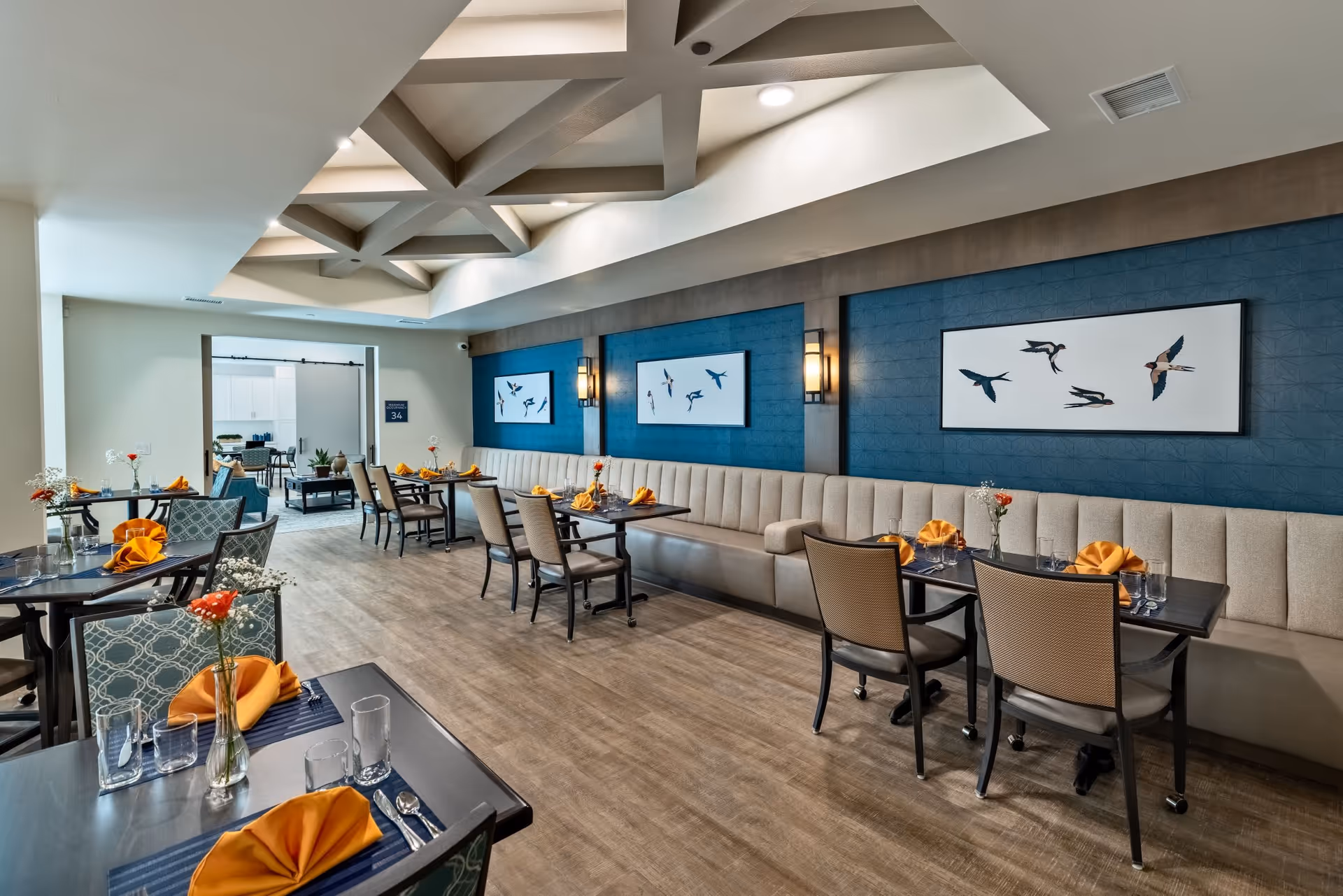 A modern dining room with several tables set with orange napkins, glasses, and silverware. The room features a long beige cushioned bench along a blue accent wall decorated with framed artwork of birds in flight. The ceiling has a geometric design with recessed lighting, and the floor is wood. In the background, there is an open doorway leading to another room with additional seating.